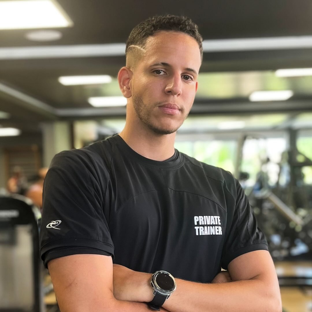 Caio Ernani de braços cruzados na academia, usando camiseta de personal trainer